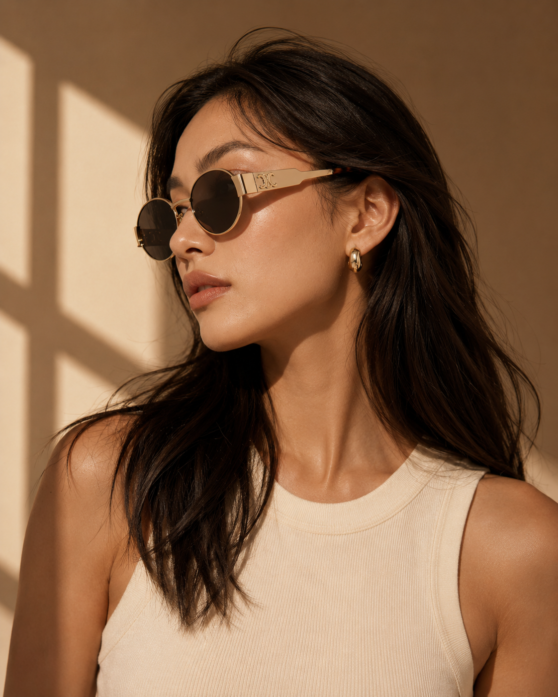 A woman with long dark hair models round sunglasses with gold metal frames and dark lenses, wearing a cream ribbed tank top against a warm beige background with soft window shadows.