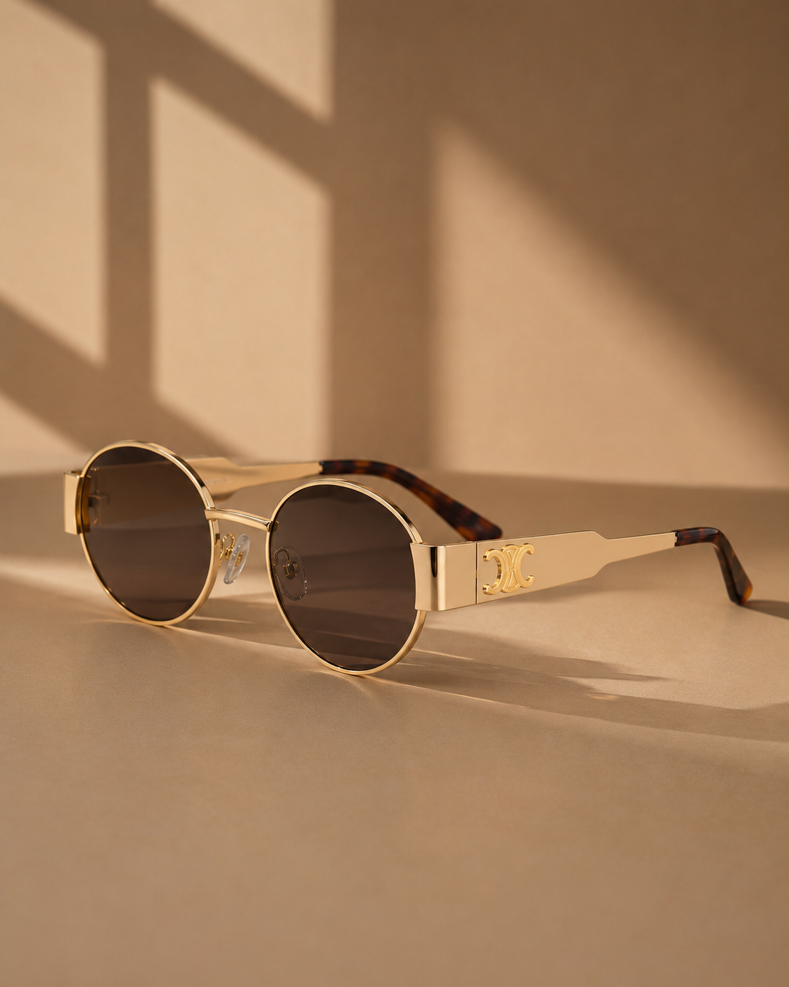 Oval sunglasses with a gold-tone metal frame, dark tinted lenses, and gold-tone bridge details, resting on a beige surface under warm window shadows.