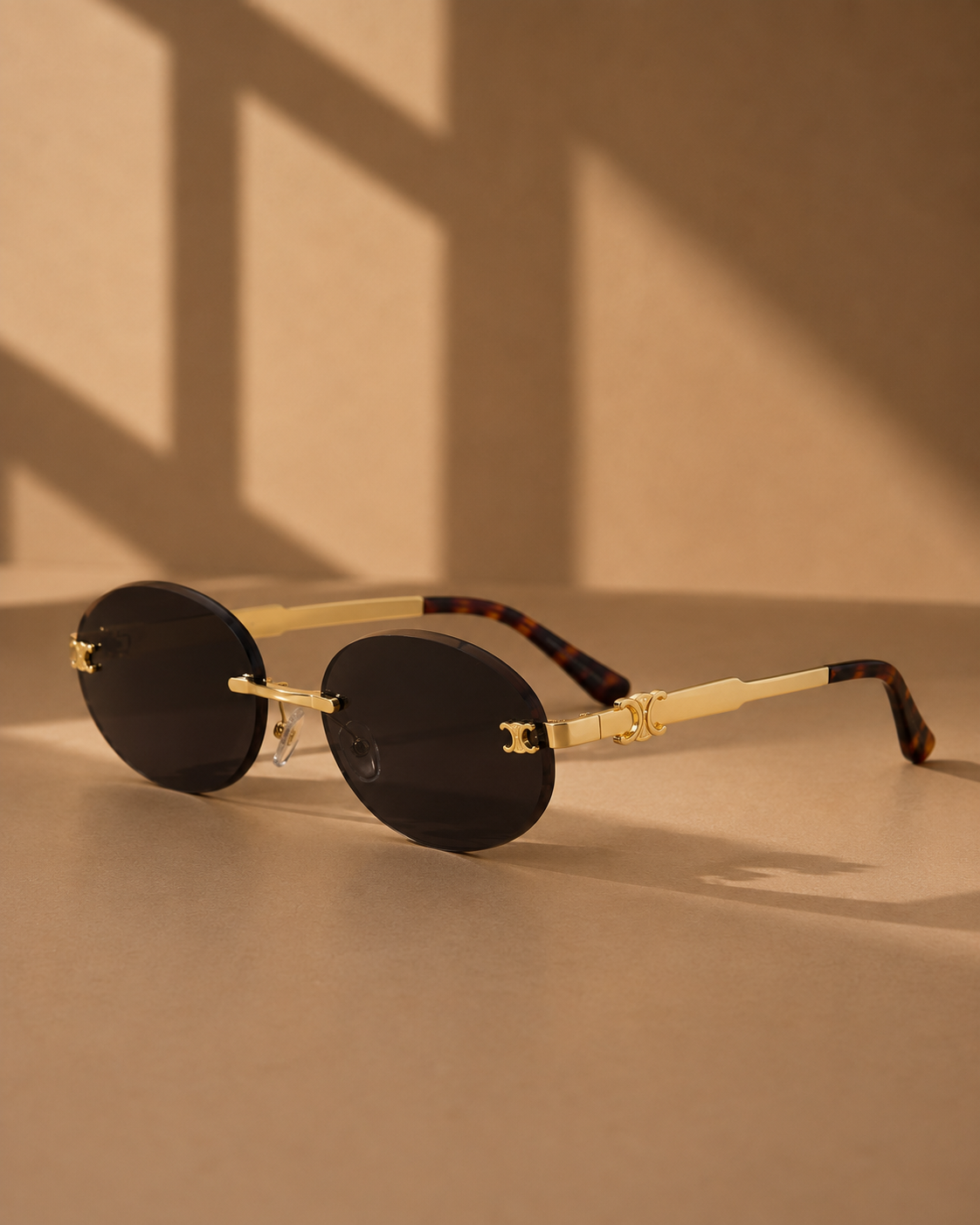 Oval sunglasses with dark lenses, gold metal frames, and tortoiseshell temples, displayed on a beige surface with warm shadow lighting.